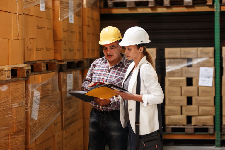 Mujer gerente y trabajador de almacén revisando listas de inventario y cumplimiento de normas de seguridad en un centro de distribución logístico.