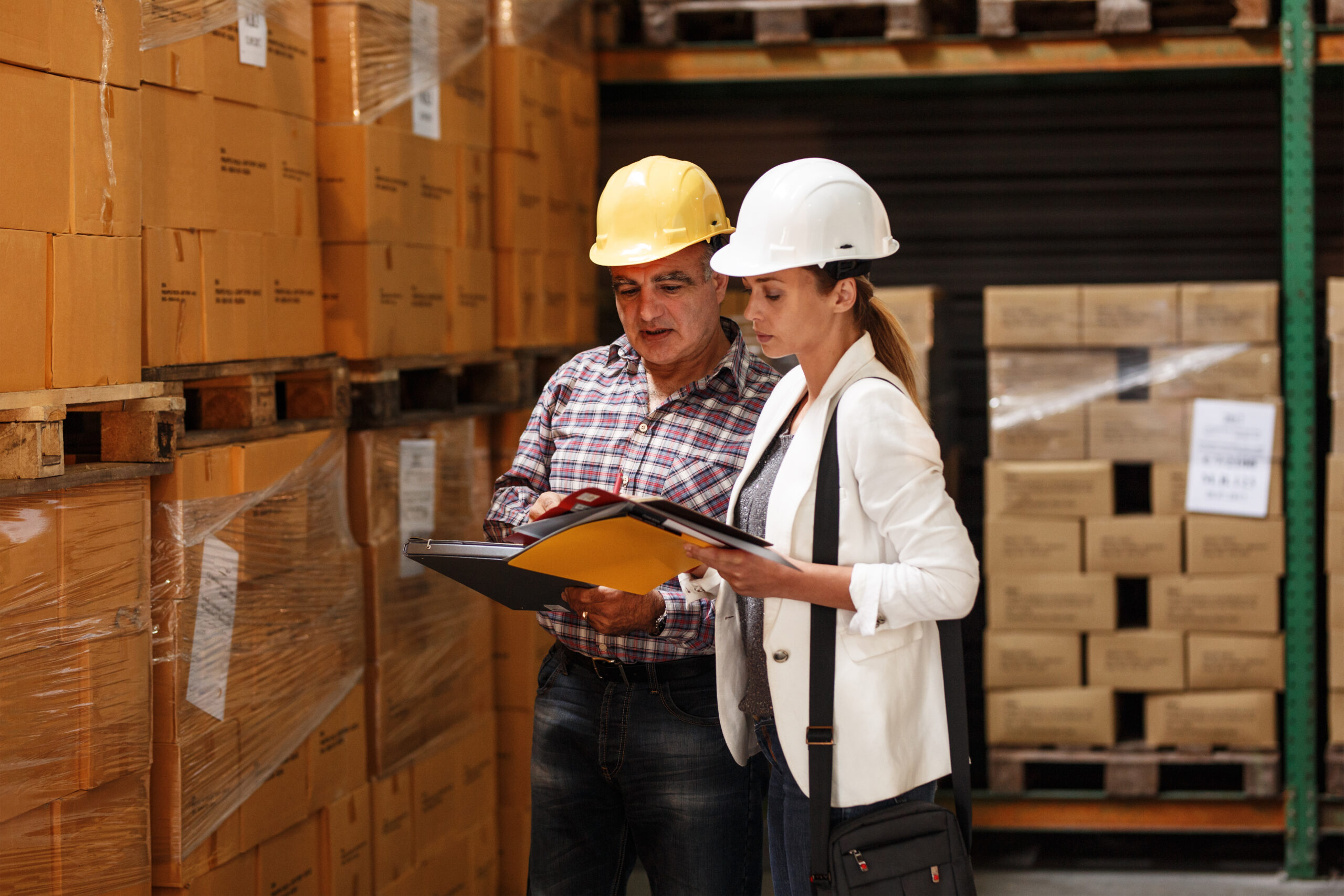 Mujer gerente y trabajador de almacén revisando listas de inventario y cumplimiento de normas de seguridad en un centro de distribución logístico.